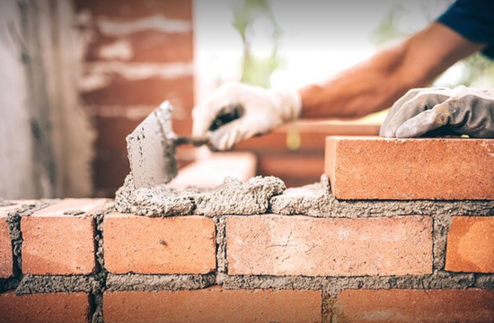 Worker laying bricks with mortar on a wall, hands visible in gloves, outdoors.