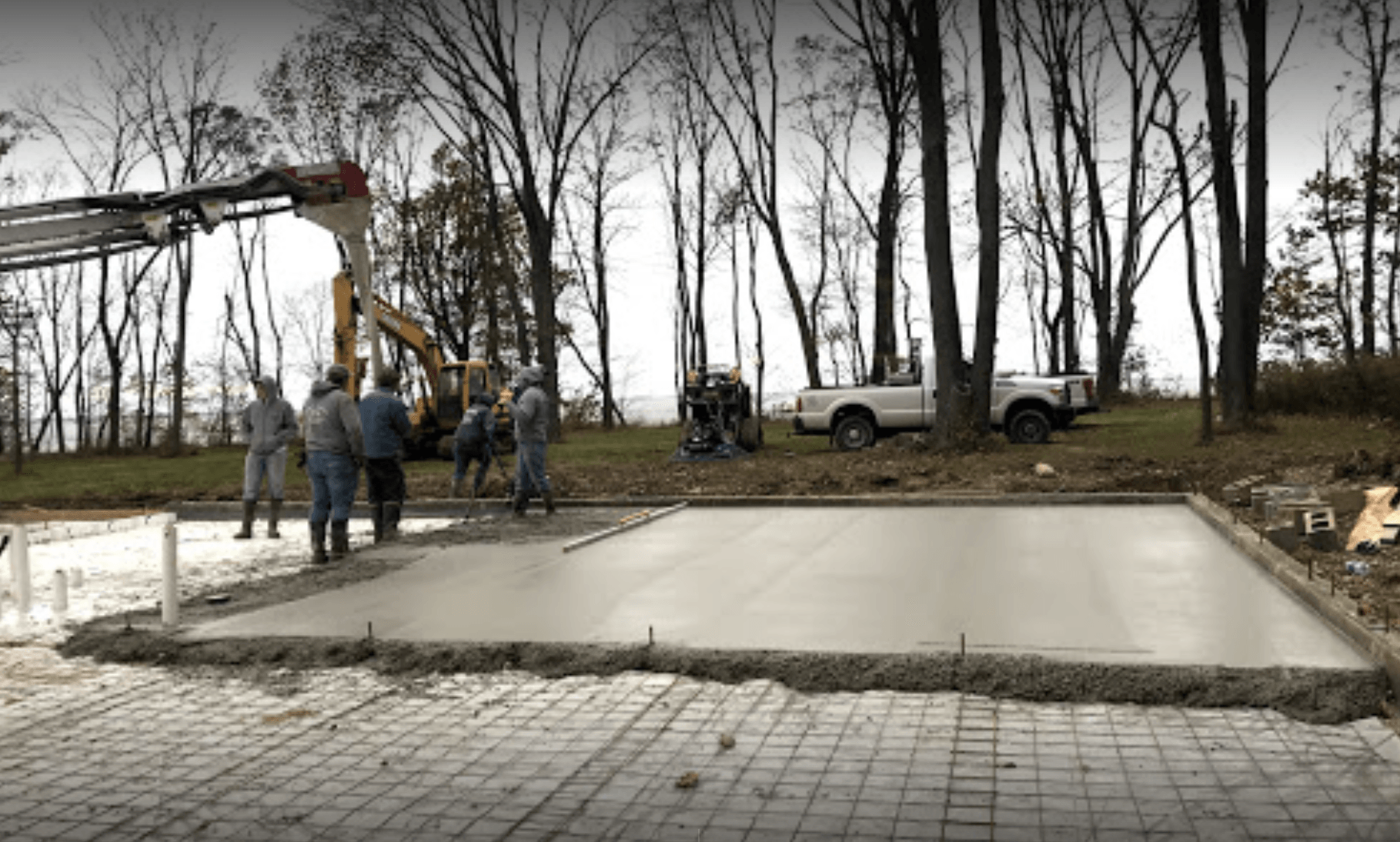 Workers pour concrete for a new driveway beside bare trees and parked trucks.