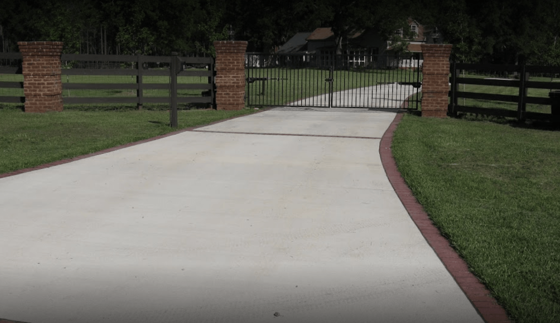 Concrete driveway leading to a closed black gate between brick pillars and wooden fences