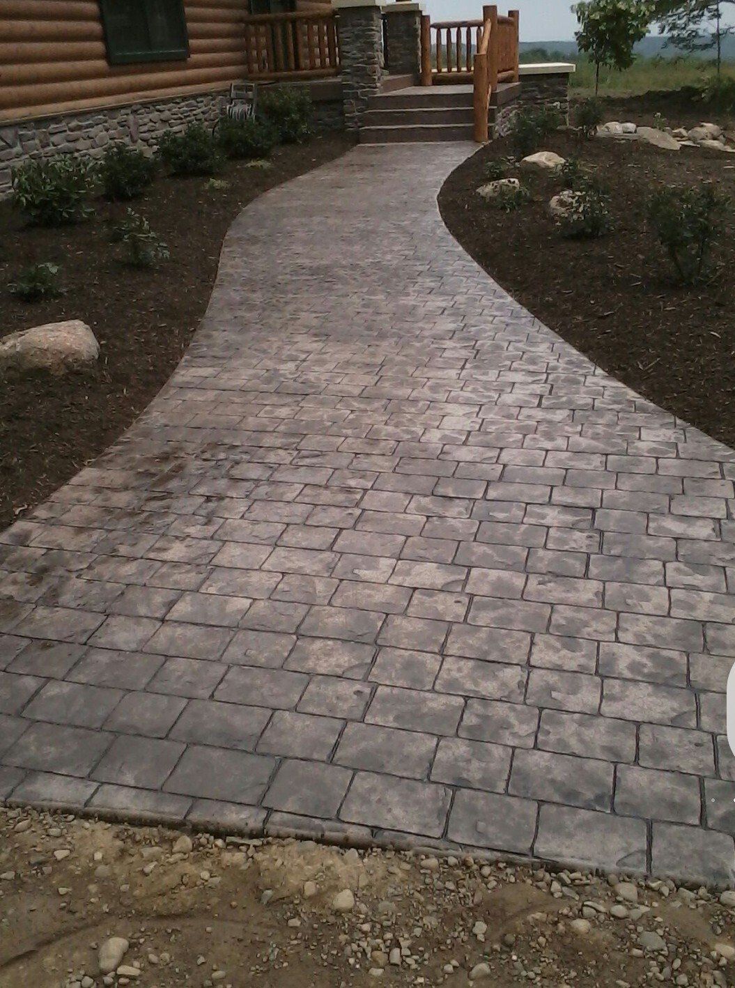 Stone walkway leading to a log cabin entrance, bordered by landscaped shrubs and rocks.