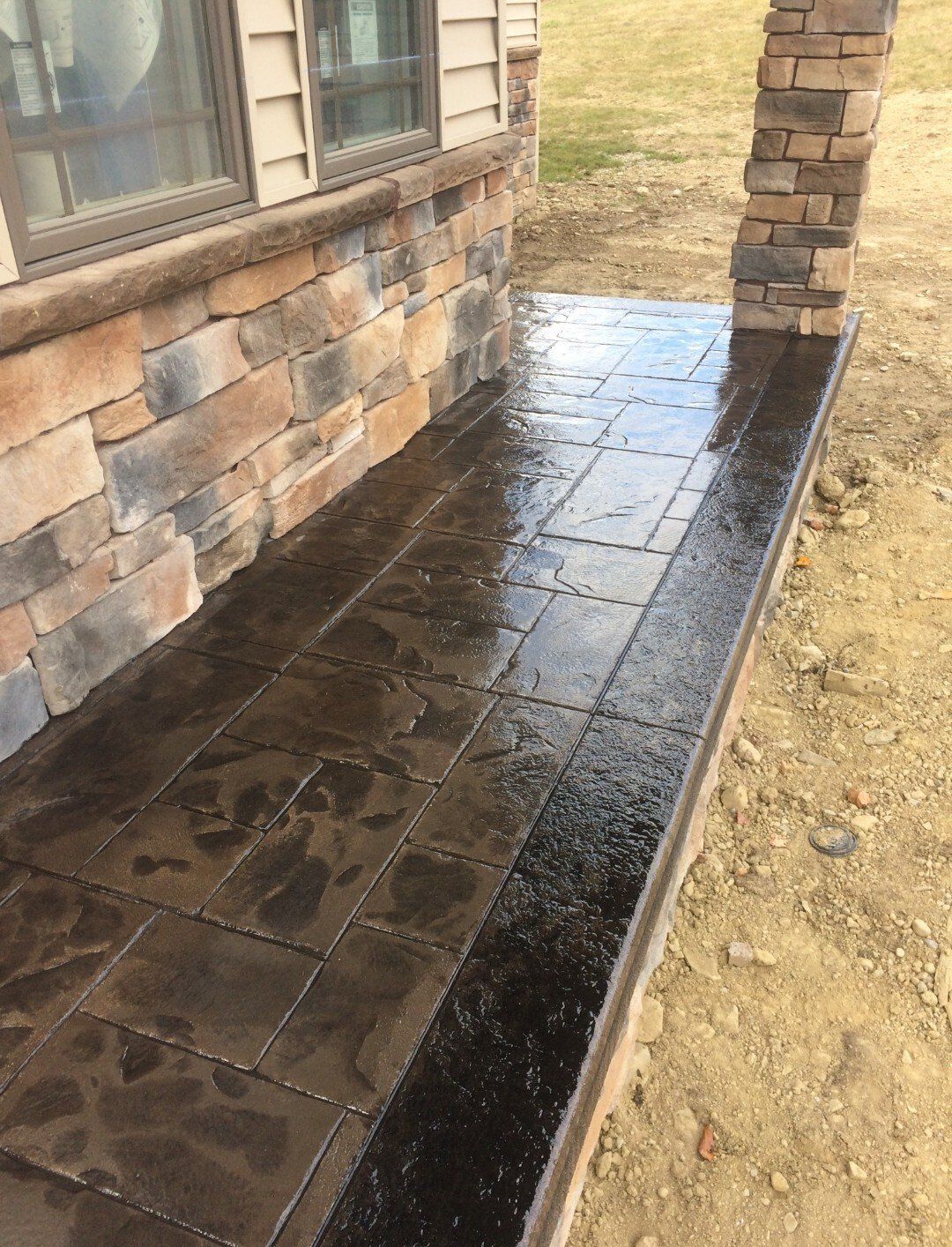 Wet dark stone patio beside a brick house and pillar, with a raised edge and dirt yard