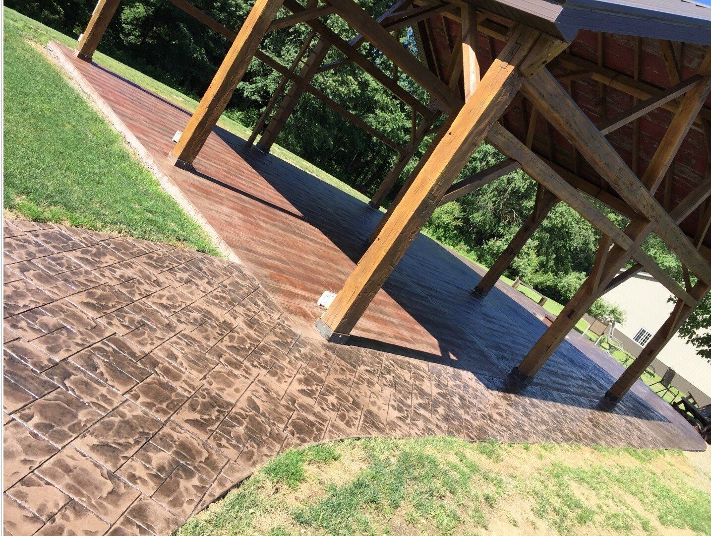 Tilted view of a wooden pavilion with yellow posts over a brick path and green grass, beside a pool.